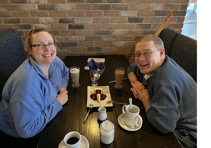 Yolanda, wearing glasses and a light blue sweater, and James, wearing a dark grey sweater, sitting at a booth at a restaurant, with dessert and drinks on the table.